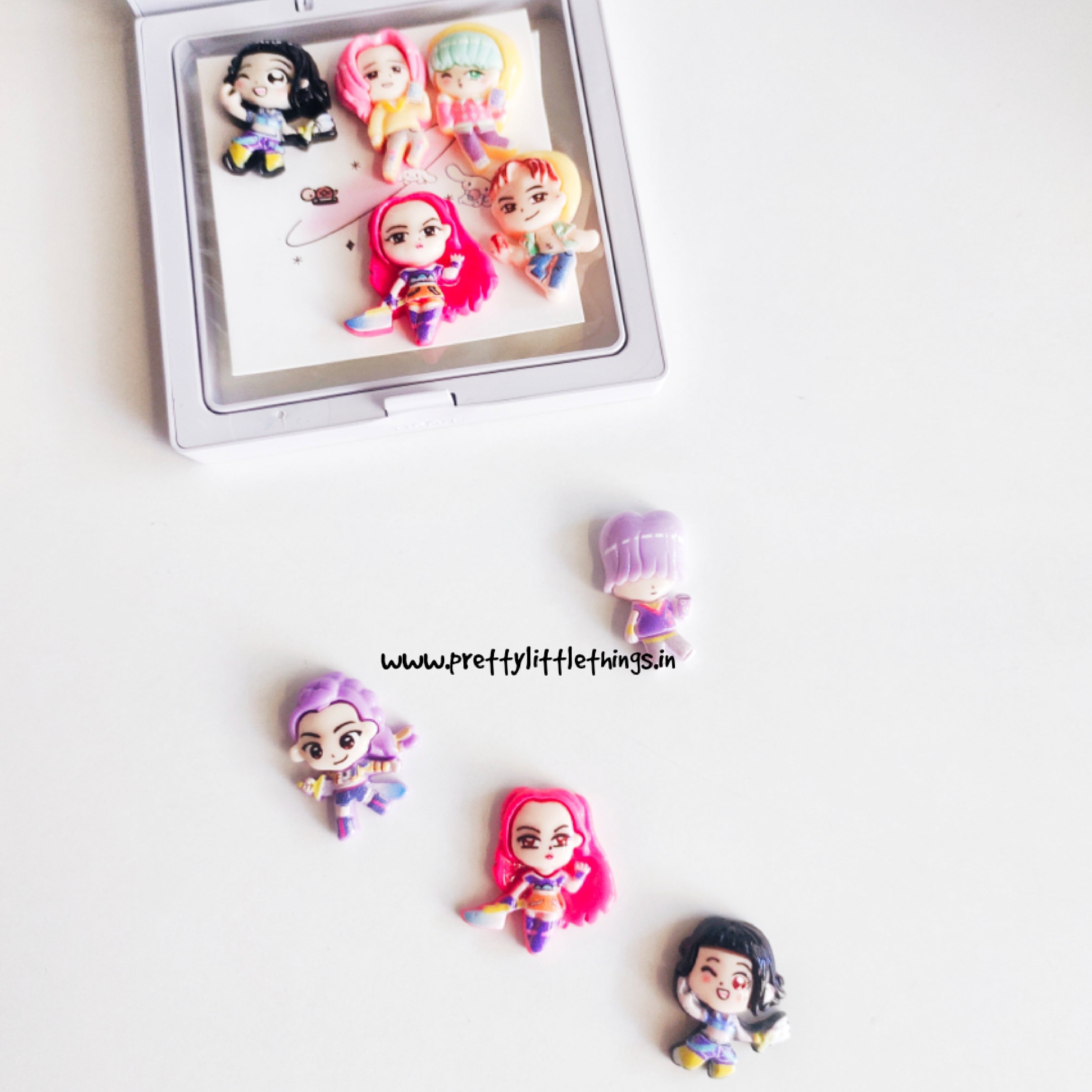 Small figurines of k pop demon hunters characters on a white surface with a branded watermark.