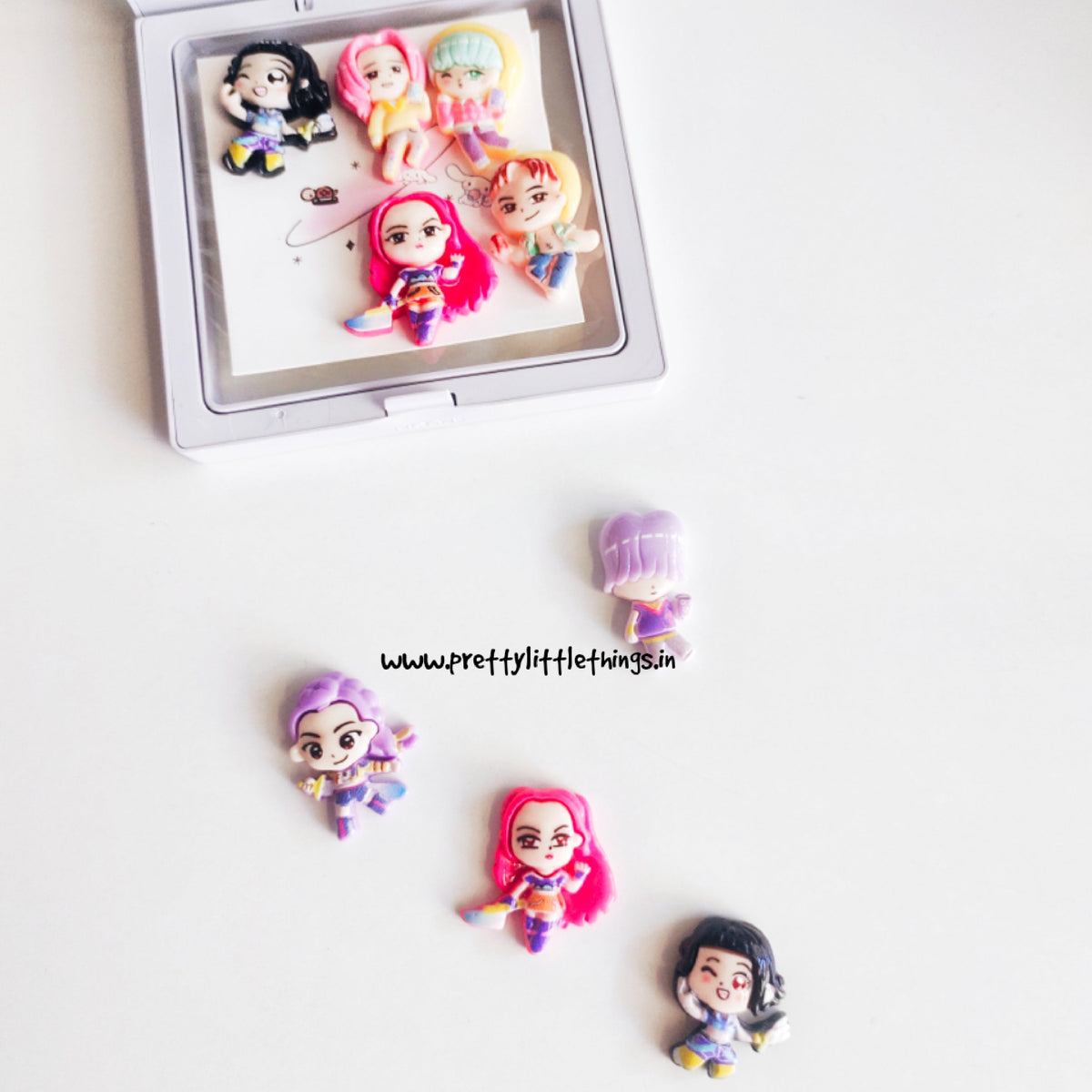 Small figurines of k pop demon hunters characters on a white surface with a branded watermark.